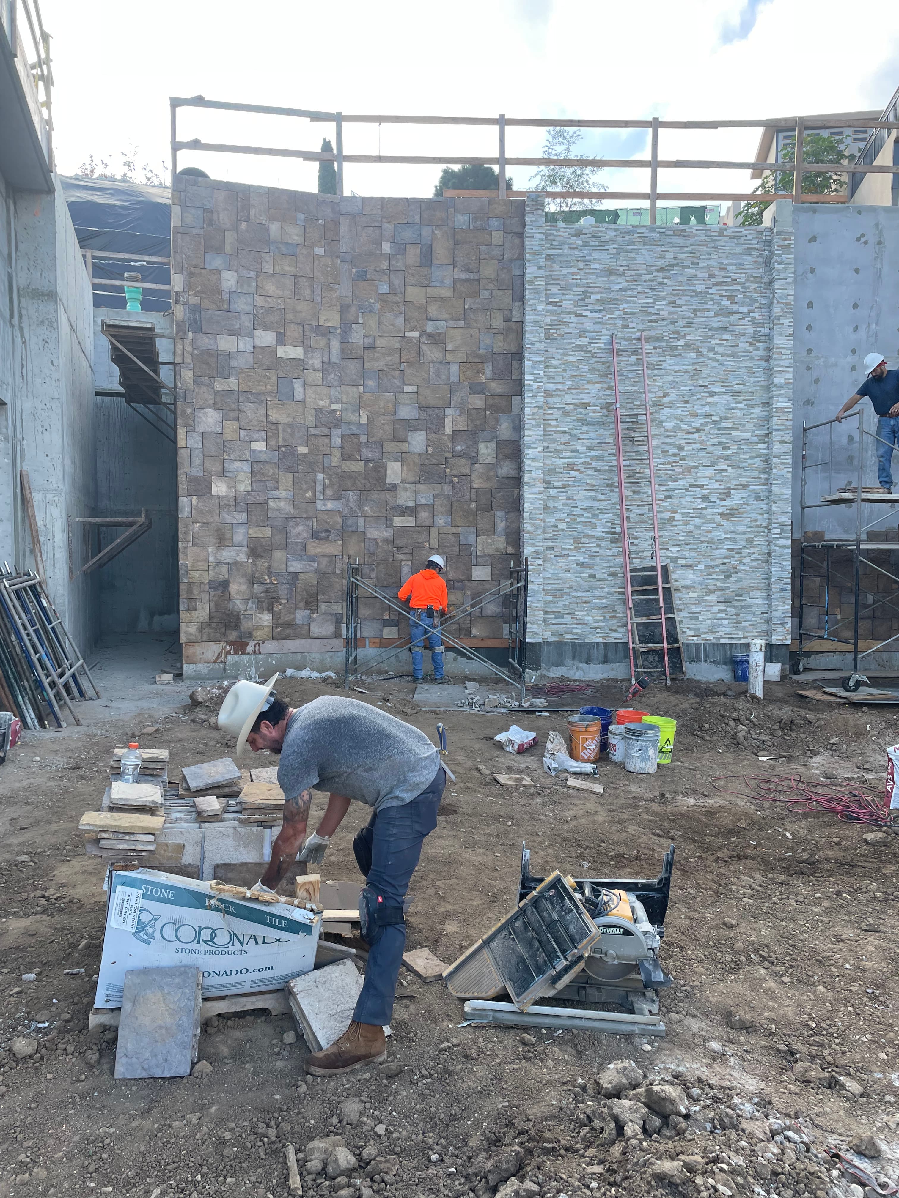Construction workers installing stone veneer on large exterior walls at a dusty building site.