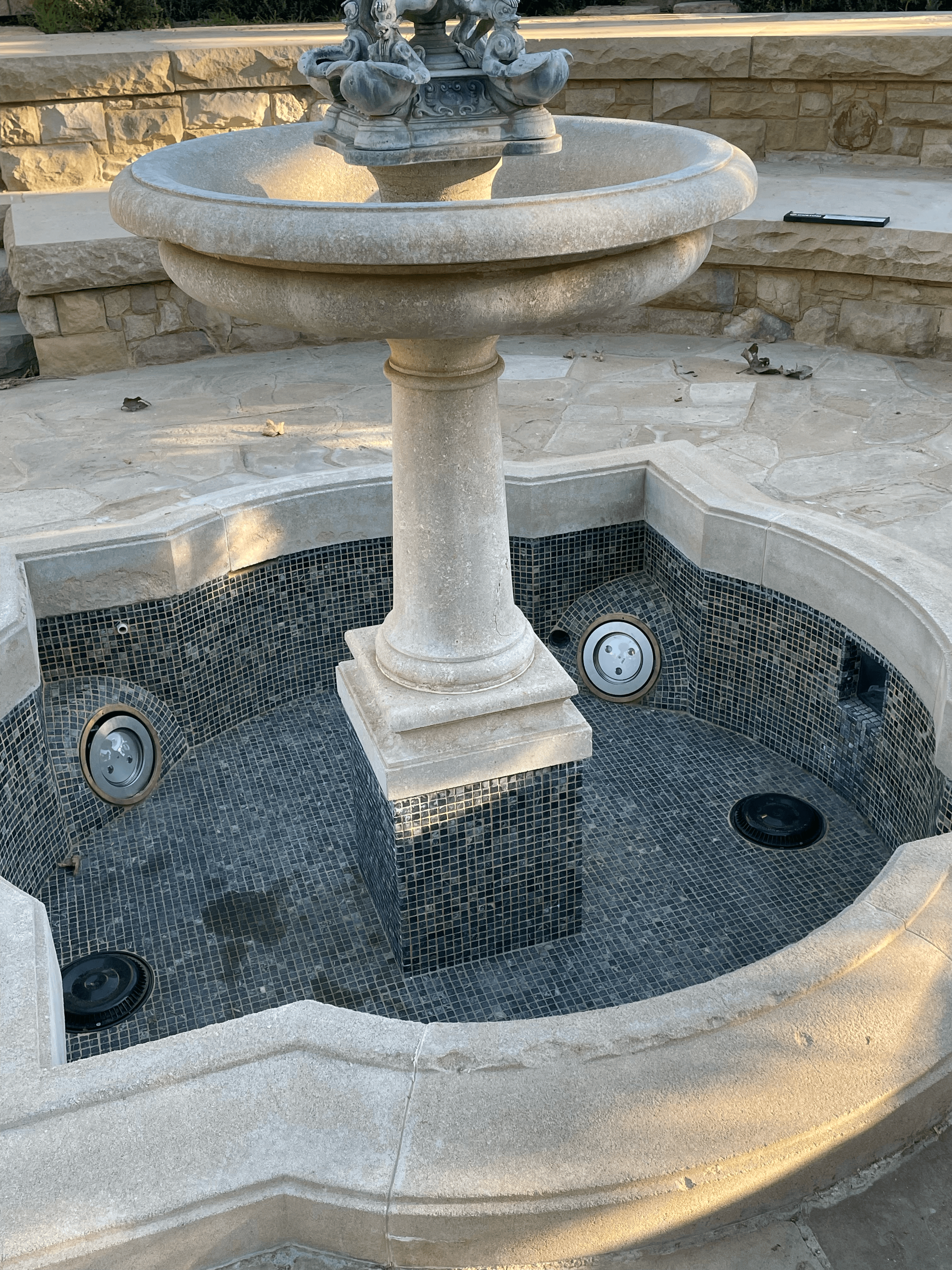 Empty tiered stone fountain with a dark tiled basin and decorative sculpture on top.